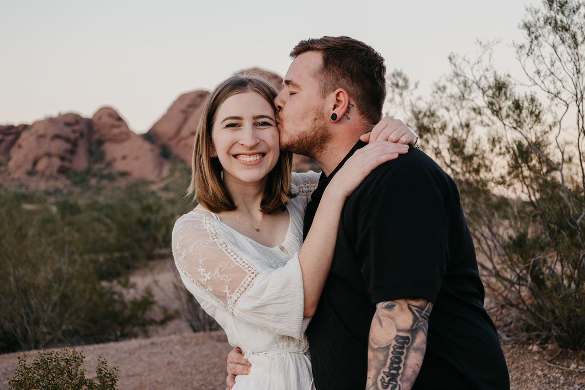 Man kissing his girlfriend on the forehead while she looks at the camera.