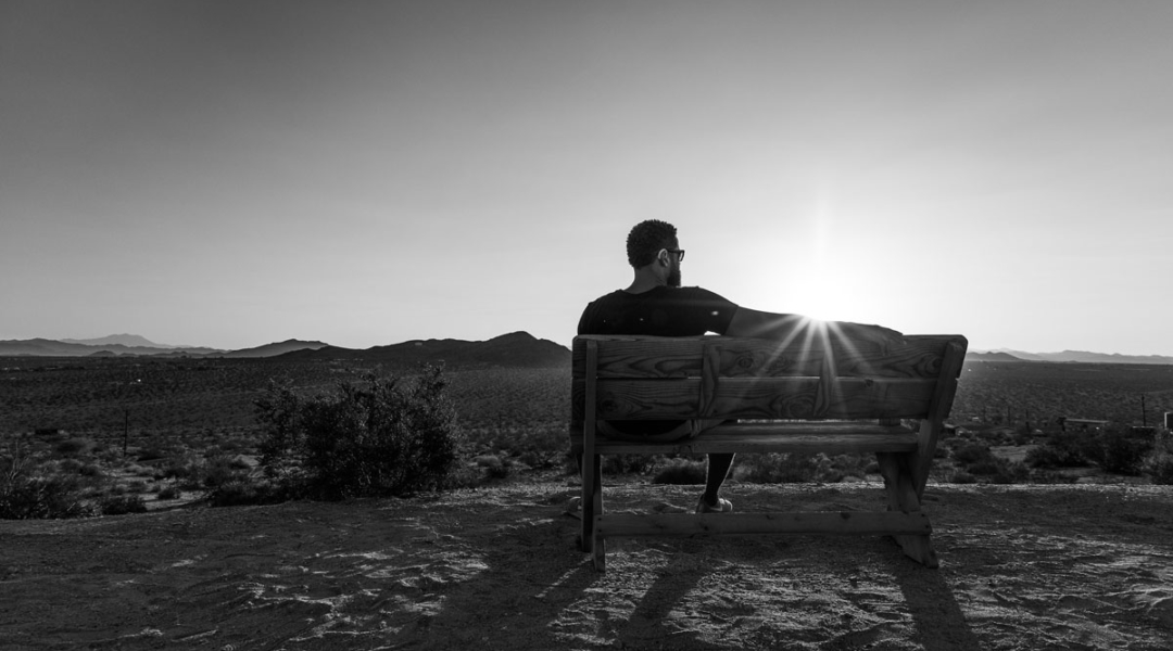 Black and white photograph of a man sitting on a bench looking out to the desert landscape.