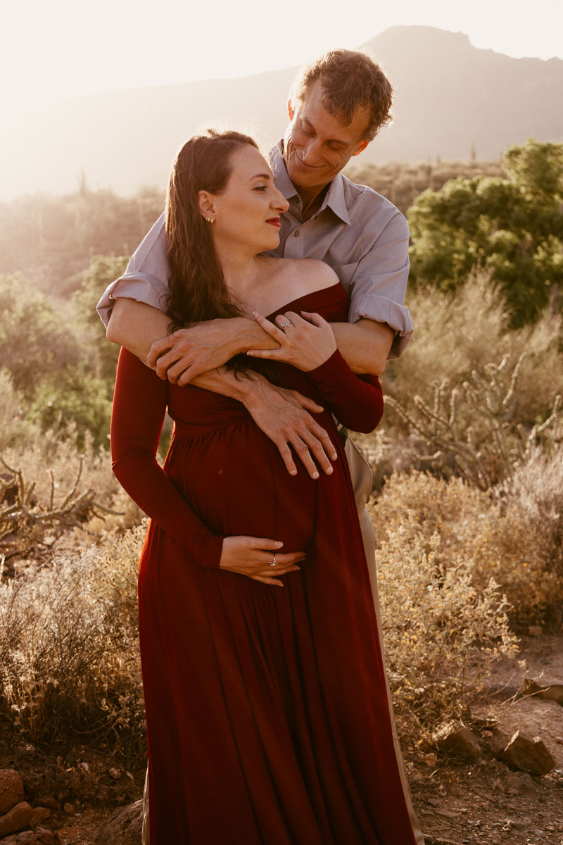 Tall man hugging a pregnant woman from behind with a desert background.